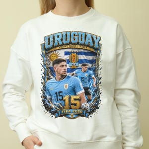 May include: A cream-colored crewneck sweatshirt featuring a graphic print of two soccer players in light blue Uruguay jerseys. The design includes the text URUGUAY, VISION MAGIC LEADER, and FIFA 2026, along with the Uruguayan flag, a sun emblem, and the number 15.