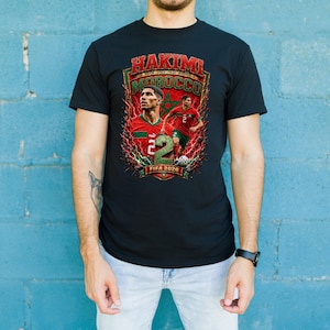 May include: A black t-shirt featuring a graphic print of soccer player Achraf Hakimi in a red and green Morocco jersey. The design includes the text HAKIMI, MOROCCO, and FIFA 2026, along with a large green number 2 and a star emblem background.