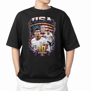 May include: A black t-shirt featuring a graphic print of a soccer player in a white jersey with the number 10. The design includes the text USA, VISION | MAGIC | LEADER, and FIFA 2026, set against a background of the American flag and lightning.