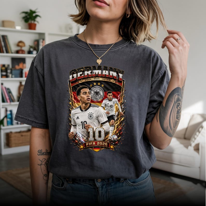 May include: A charcoal gray short-sleeve t-shirt featuring a graphic print of a soccer player in a white jersey with the number 10. The design includes the text GERMANY, VISION, MAGIC, CAPTAIN, and FIFA 2026, along with the German flag colors.