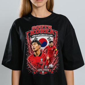 May include: A black t-shirt featuring a graphic print of a soccer player in a red jersey. The design includes the text SOUTH KOREA, FIFA 2026, and VISION | SPEED | PRIDE | KOREA, along with a red and blue circular emblem and red lightning effects.