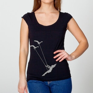Op de afbeelding: Een zwart t-shirt met ronde hals en een grijze afbeelding van een meisje dat op een schommel schommelt met vogels die erboven vliegen.