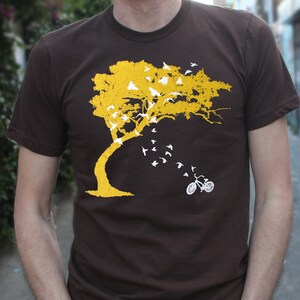 May include: A brown t-shirt with a yellow tree and white birds flying away from it. A white bicycle is at the bottom of the tree.