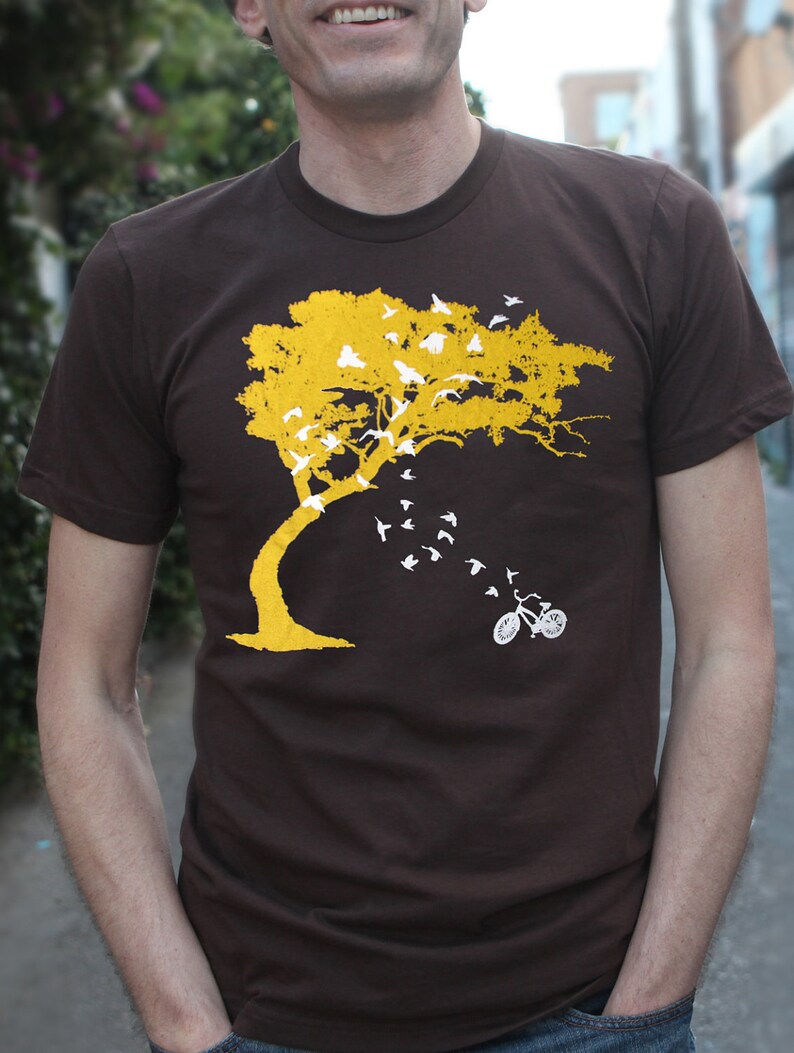 May include: A brown t-shirt with a yellow tree and white birds flying away from it. A white bicycle is at the bottom of the tree.