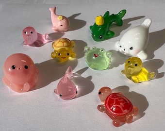 May include: A selection of small, brightly coloured sea creature figurines. The collection includes a pink octopus, a pink whale, a green dolphin, a white seal, a yellow duck, a yellow turtle, a green crab, and a red turtle. These are likely resin or similar.