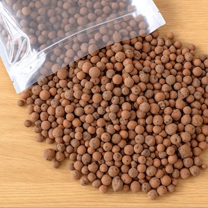 May include: A clear plastic bag partially open, spilling out a pile of brown, round clay pebbles. The pebbles vary in size and are scattered on a light brown wooden surface. The image is well-lit, highlighting the texture of the clay.