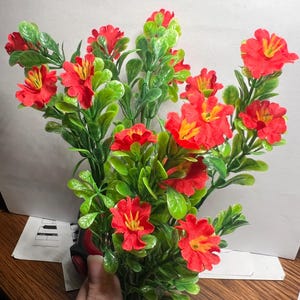 Op de afbeelding: Een levendig boeket kunstbloemen met helderrode bloesems met gele centra en weelderige groene bladeren. Het arrangement is een mix van kleine en grote bloemen, waardoor een volle en getextureerde uitstraling ontstaat. Perfect voor woondecoratie of knutselen.