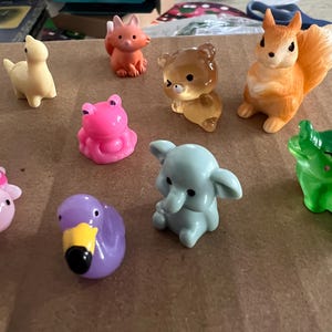 Puede incluir: Una colección de pequeñas figuras de animales coloridas. El surtido incluye un dinosaurio amarillo, un zorro naranja, un oso transparente, una ardilla naranja, una rana rosa, un elefante azul, un caimán verde, una tortuga rosa y un pato morado.