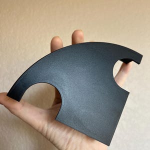 May include: A black, curved object with a textured surface and two circular cutouts. The object is held in a hand, showcasing its shape and design. The item appears to be a decorative or functional piece.