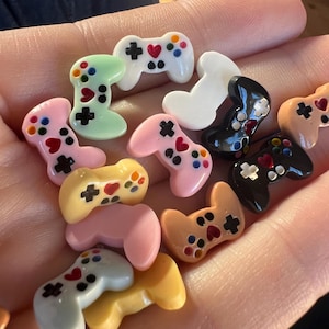 Puede incluir: Una colección de mini mandos de juego coloridos en varios tonos, incluyendo rosa, verde, amarillo y negro. Cada mando presenta un diseño con botones de cruz, corazón y puntos. Los mandos son pequeños y parecen estar hechos de un material brillante.