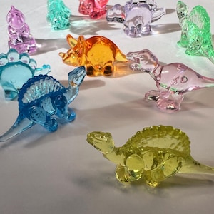 May include: A collection of colorful, transparent dinosaur figurines in various shapes and sizes. The dinosaurs are in shades of blue, pink, yellow, orange, and green. The figurines are made of a clear, glossy material.