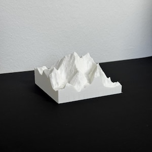 Puede incluir: Modelo de cordillera impreso en 3D en blanco. El modelo presenta picos y valles detallados, mostrando una representación realista de un paisaje montañoso. La base es rectangular y el diseño es minimalista.
