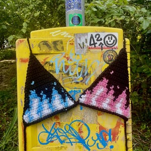 May include: A black crochet bikini top with blue and white flame-like patterns on one cup and pink and white on the other. The bikini is displayed on a yellow surface covered in graffiti and stickers, with a green background.