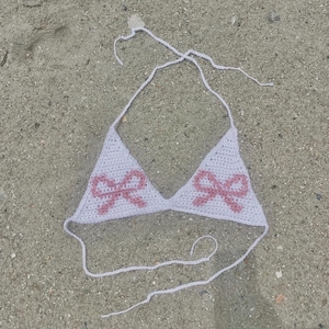 May include: White crochet bikini top with pink bow detailing. The triangle-shaped top has a halter neck and tie-back design. The top is lying on a sandy surface, ideal for beach or poolside wear.