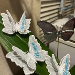 May include: Three white embroidered butterfly patches with light blue cursive text reading Grandpa, Bampy, and Grandma, alongside one stained glass butterfly ornament with dark purple and grey wings mounted on a thin gold metal wire stem.
