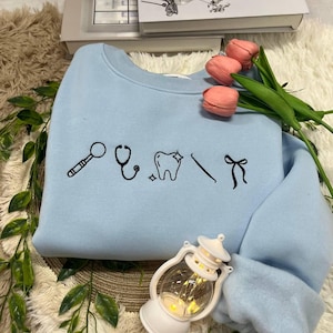 Puede incluir: Una sudadera azul claro con dibujos lineales negros de instrumentos dentales y un lazo. El diseño incluye un espejo dental, un estetoscopio, un diente, un explorador dental y un lazo. La sudadera se muestra con flores rosas y una linterna decorativa.