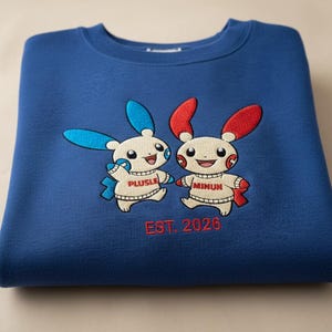 May include: A folded blue sweatshirt featuring embroidered cartoon characters, one with blue ears and the other with red ears. The characters are labeled "Plusle" and "Minun" with the text "EST. 2026" in red below.
