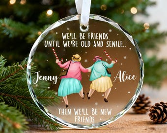 May include: A clear, round Christmas ornament with a white ribbon. The ornament features an illustration of two elderly women holding hands, with the text "WE'LL BE FRIENDS UNTIL WE'RE OLD AND SENILE.. THEN WE'LL BE NEW FRIENDS."
