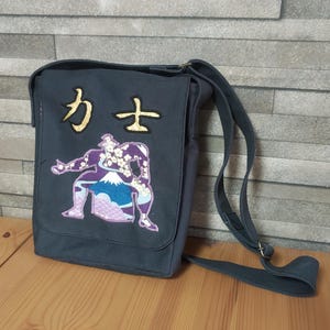 以下が含まれることがあります： 富士山と桜の模様が刺繍された力士のデザインが施されたダークグレーのキャンバス地メッセンジャーバッグ。力士の図案の上には金色の日本語の漢字が2文字刺繍されています。バッグには調節可能なショルダーストラップが付いています。