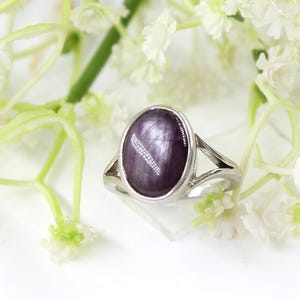 Peut inclure: Une bague en argent ornée d'une pierre ovale de couleur violet foncé. La bague est posée sur un fond de fleurs blanches et de tiges vertes, créant une esthétique naturelle et élégante. La pierre précieuse a une qualité réfléchissante subtile.