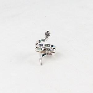 Peut inclure: Bague serpent argentée ornée d'incrustations nacrées iridescentes. La bague s'enroule autour du doigt, avec la tête du serpent en haut et la queue courbée en bas. La bague est posée sur un fond blanc.
