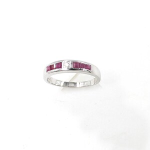 May include: A silver ring featuring a row of five square-cut, deep red gemstones, with a single, clear, square-cut gemstone set between them. The ring has a smooth, polished band and a classic design.