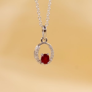 Peut inclure: Un collier pendentif en argent avec une petite pierre précieuse rouge foncé de forme ovale au centre. La pierre précieuse est entourée d'un halo de petites pierres claires. Le pendentif est suspendu à une fine chaîne en argent.