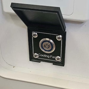 May include: A black rectangular panel with a hinged lid, mounted on a white surface. The panel features a silver button with a power symbol and the text "Cooling Fan" below it. Four silver screws secure the panel.