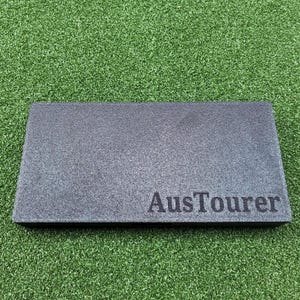 May include: A rectangular, dark grey object with a textured surface. The word "AusTourer" is printed in a darker shade on the bottom right corner. The object is set against a green, textured background.
