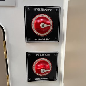 May include: Two square black electrical switch panels mounted on a white surface. Each panel features a red circular rotary switch with an 'm' logo, labelled 'INVERTER+LOAD' and 'BATTERY MAIN' respectively, with the brand name 'EZYTRAIL' printed below.