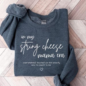 May include: A dark gray sweatshirt with the text "in my string cheese mama era" in white script. Below the text is "overwhelmed touched out but exactly who I'm meant to be" and a heart symbol.