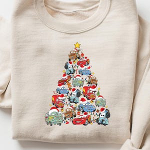 May include: A beige sweatshirt featuring a Christmas tree design made of various cartoon cars. The cars are in different colors and are wearing Santa hats. A yellow star tops the tree. The sweatshirt is folded on a white surface.