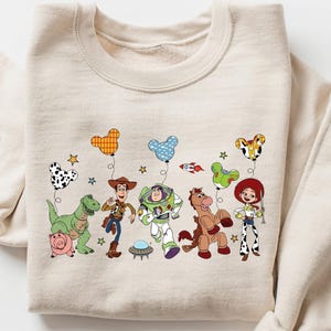 May include: A beige sweatshirt featuring a colorful graphic of animated characters and balloons. The design includes a dinosaur, a pig, a cowboy, a space ranger, a horse, and a cowgirl. The balloons have various patterns and shapes.