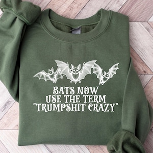 May include: An olive green sweatshirt with the phrase "BATS NOW USE THE TERM 'TRUMPSHIT CRAZY'" in white. The design features three stylized bats. The sweatshirt is folded on a light wood-grain surface.