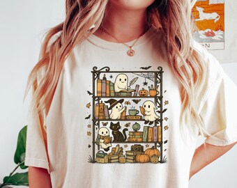 May include: Cream-colored t-shirt featuring a whimsical illustration of a bookshelf with Halloween-themed characters. The design includes ghosts, a black cat, pumpkins, and books. The shirt is a comfortable, casual style.