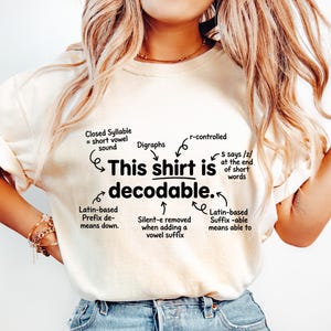 May include: A cream-colored short-sleeve t-shirt featuring black text that reads: This shirt is decodable. The design includes arrows pointing to linguistic notes: Closed Syllable, Digraphs, r-controlled, S says /z/, Latin-based Prefix de-, Silent-e, and Suffix -able.