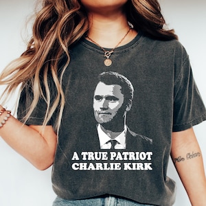 May include: Dark gray t-shirt featuring a portrait of a man in a suit and tie. The text "A TRUE PATRIOT CHARLIE KIRK" is printed in white below the portrait. The shirt is worn with blue jeans and white sneakers.