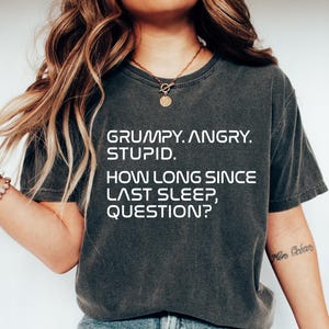 May include: Dark gray t-shirt with the text "GRUMPY. ANGRY. STUPID. HOW LONG SINCE LAST SLEEP, QUESTION?" in white. The shirt is paired with blue jeans and white sneakers. The overall theme is casual wear.