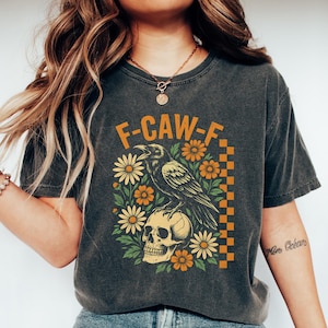 May include: Dark gray t-shirt featuring a graphic design with the text "F-CAW-F". The design includes a raven perched on a skull, surrounded by orange and white flowers. The shirt is worn with blue jeans and white shoes.