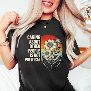 May include: Black t-shirt with the text "Caring About Other People Is Not Political" in white. The shirt features a floral design with a sunset background and hands cradling the flowers. The design is centered on the front of the shirt.