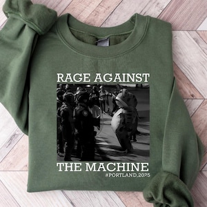 May include: An olive green sweatshirt featuring the text "RAGE AGAINST THE MACHINE" and a black and white image of a protest. The text "#PORTLAND.2025" is below the image. The sweatshirt has long sleeves and a crew neck.