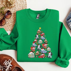 May include: A green sweatshirt featuring a Christmas tree design made of cartoon cars, each adorned with Santa hats. A yellow star tops the tree. The sweatshirt is a crew neck style.