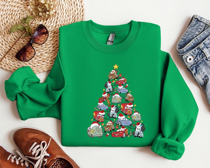 May include: A green sweatshirt featuring a Christmas tree design made of cartoon cars, each adorned with Santa hats. A yellow star tops the tree. The sweatshirt is a crew neck style.