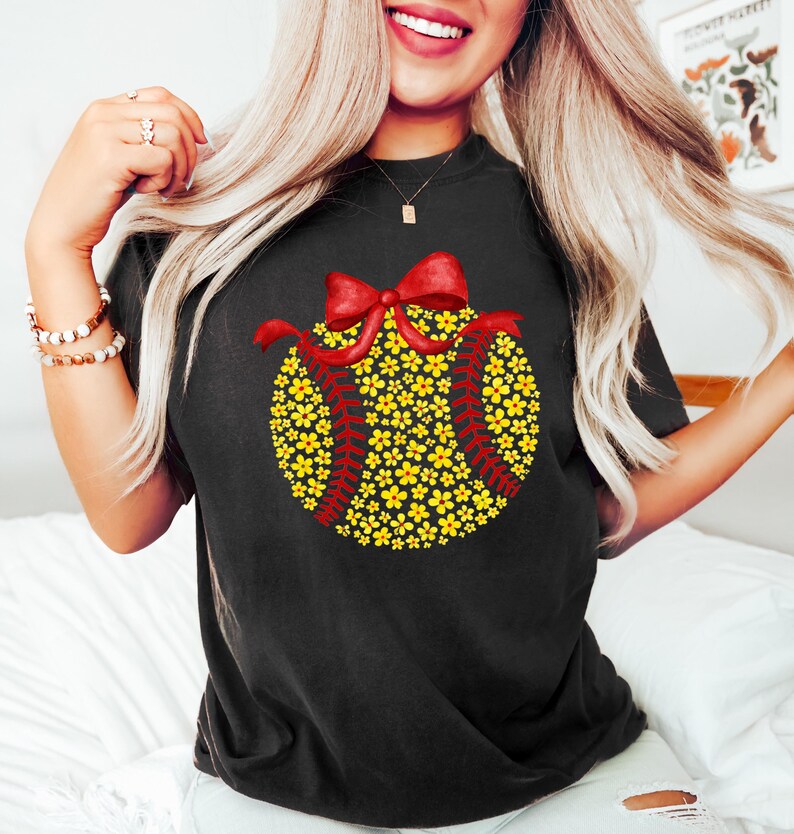 May include: Black t-shirt featuring a softball design. The softball is composed of yellow flowers and red stitching, topped with a red bow. The shirt is a comfortable fit, perfect for casual wear or sports events.