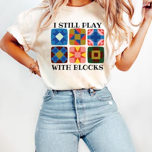 May include: Cream-colored t-shirt with the text "I STILL PLAY WITH BLOCKS" above a grid of six colorful quilt block designs. The blocks feature various geometric patterns in shades of blue, red, yellow, and green. The shirt is paired with light blue jeans.