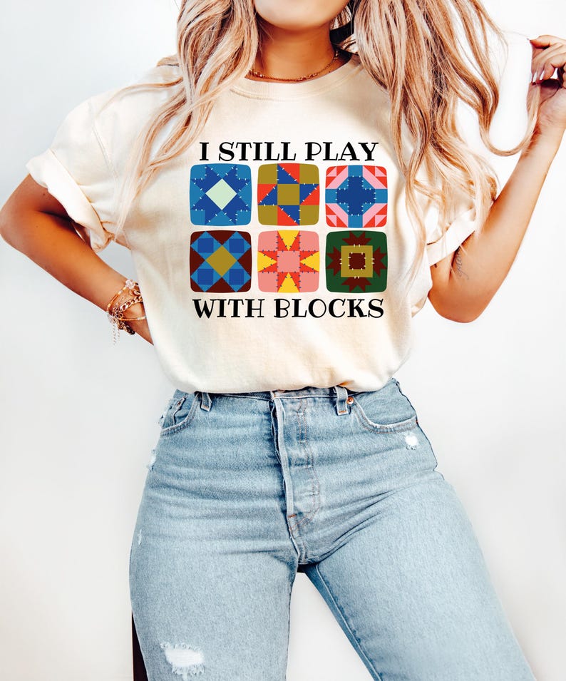 May include: Cream-colored t-shirt with the text "I STILL PLAY WITH BLOCKS" above a grid of six colorful quilt block designs. The blocks feature various geometric patterns in shades of blue, red, yellow, and green. The shirt is paired with light blue jeans.
