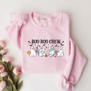 May include: A pale pink sweatshirt with the words "BOO BOO CREW" in black. The design features cartoon ghosts dressed as medical staff, with bats and stars. The sweatshirt has a crew neck.