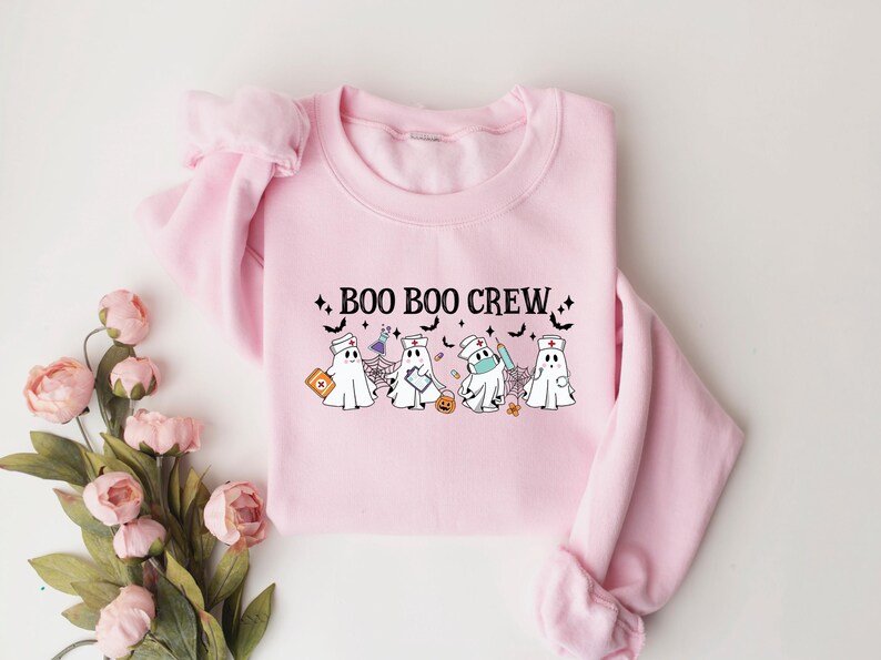 May include: A pale pink sweatshirt with the words "BOO BOO CREW" in black. The design features cartoon ghosts dressed as medical staff, with bats and stars. The sweatshirt has a crew neck.