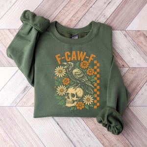 May include: Green sweatshirt with the text "F-CAW-F" above a graphic of a raven perched on a skull, surrounded by flowers. The design includes orange and yellow accents and a checkered border.