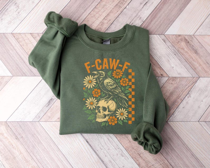 May include: Green sweatshirt with the text "F-CAW-F" above a graphic of a raven perched on a skull, surrounded by flowers. The design includes orange and yellow accents and a checkered border.